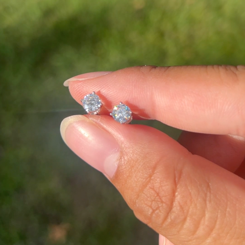 A close-up of a person's fingers holding a pair of round-cut diamond stud earrings against a blurred green outdoor background.