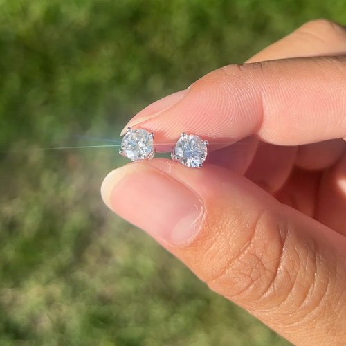 A close-up of a person's hand holding two round-cut diamond stud earrings against a blurred green outdoor background.